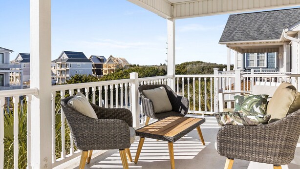 A covered outdoor seating area with wicker furniture and a wooden table, overlooking a residential neighborhood. - Family Beach Home ~Steps to Beach~Hot Tub~Game Room~Sleeps 13! (Surf City)