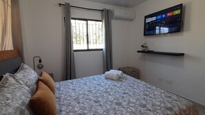 Una camera da letto con un letto, una TV fissata al muro, una finestra con tende e un piccolo ripiano con una pianta.