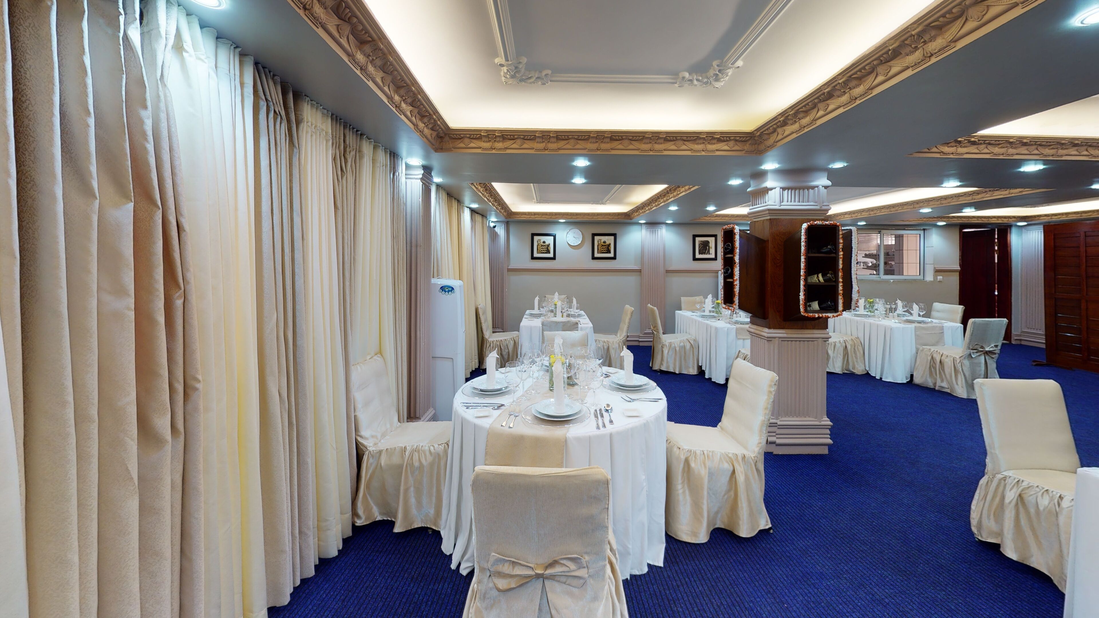A banquet hall with round tables set for a formal event, featuring white tablecloths, chairs with bows, and a blue carpeted floor.