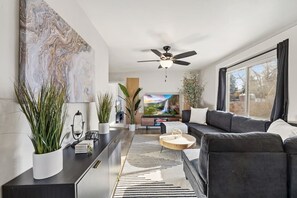 A modern living room with a large TV, dark sofas, a ceiling fan, and large windows.