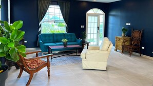 Un salón espacioso con un sofá azul, un sillón crema y una silla de madera. Hay una mesa de centro con una planta, y un ventanal con cortinas.