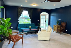 A spacious living room with a blue sofa, a cream armchair, and a wooden chair. There is a coffee table with a plant, and a large window with curtains.