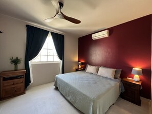 Et soveværelse med en seng, en loftsvifte, et vindue med gardiner, et natbord med en lampe og en aircondition.