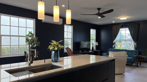 A modern kitchen with a large island, a ceiling fan, and a view of the outdoors through large windows.