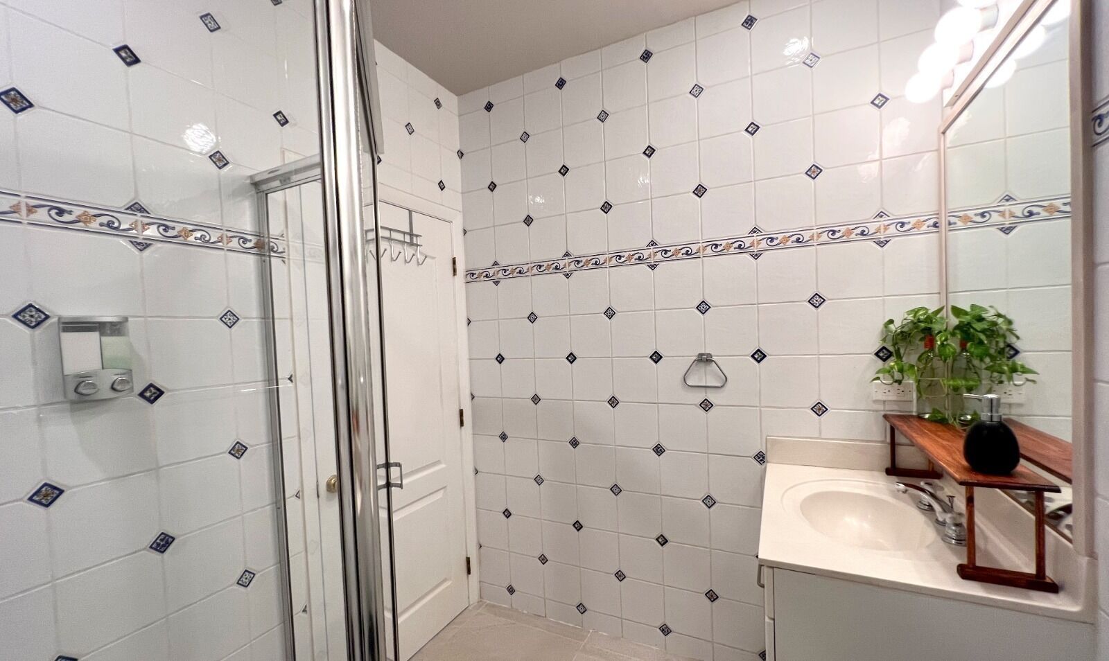 A bathroom with white tiled walls, a glass shower enclosure, a sink with a wooden vanity, and a potted plant.