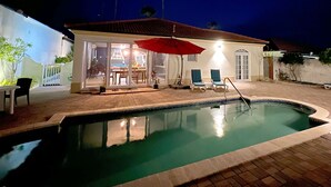 Una piscina con borde de azulejos, una sombrilla roja y una zona de patio con sillas y una mesa.