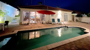 Une piscine dont le bord est recouvert de carrelage, un parasol rouge et un espace extérieur aménagé avec des chaises et une table.