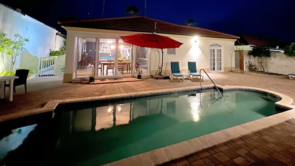 A swimming pool with a tiled edge, a red umbrella, and a patio area with chairs and a table.