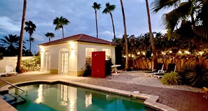 A pool area with a small building, palm trees, and outdoor seating.