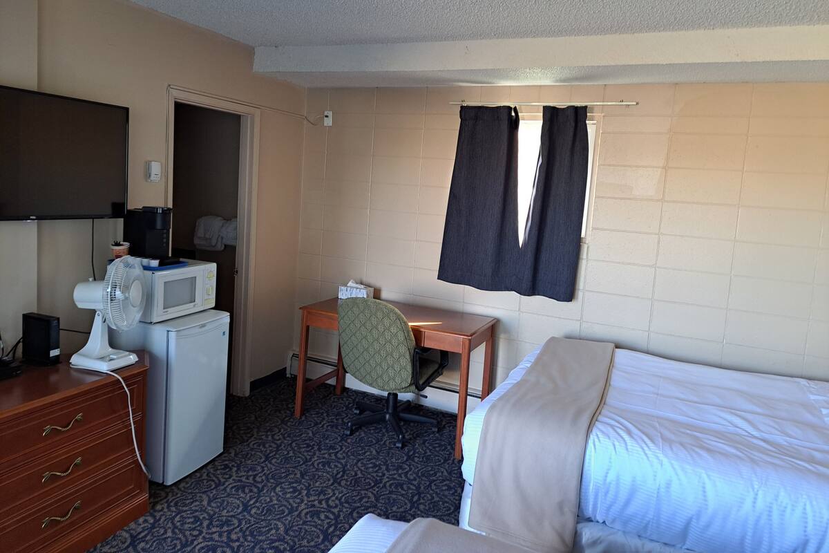 A hotel room with a bed, a desk with a chair, a microwave, a fan, and a television.