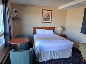 A hotel room with a bed, a bedside table with a lamp, a chair, a small round table, a wall-mounted air conditioner, and a framed picture on the wall. - Matador Motel (Drayton Valley)