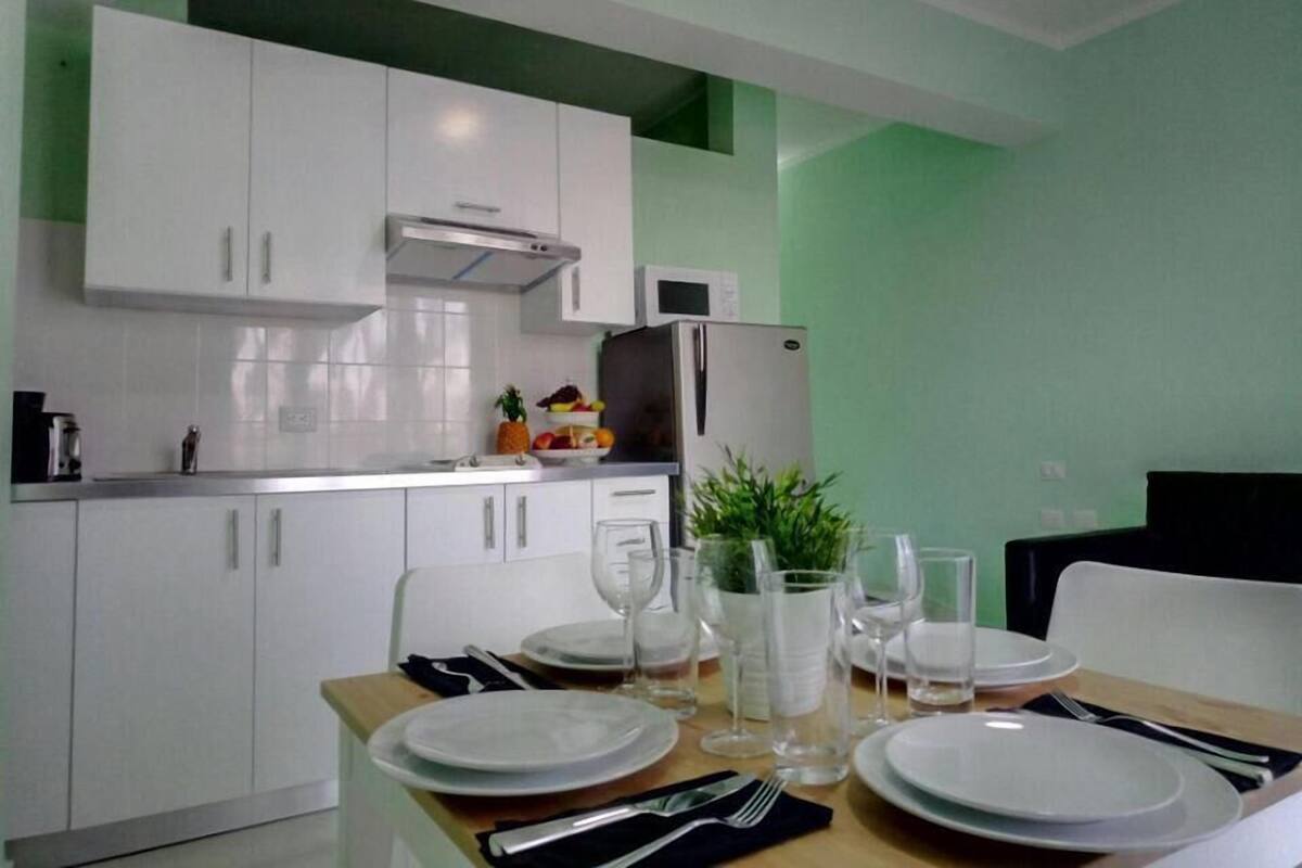 Una cocina moderna con armarios blancos, un refrigerador de acero inoxidable y una mesa de comedor dispuesta con platos, vasos y cubiertos.
