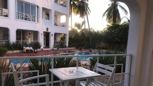 A balcony with a table and chairs overlooking a swimming pool, surrounded by palm trees and a white building. - Blue Garden Apartment Los Corales (Punta Cana)
