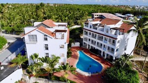 A white building with a swimming pool, surrounded by palm trees and a view of a densely vegetated area. - Blue Garden Apartment Los Corales (Punta Cana)