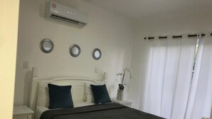 A bedroom with a bed, white nightstands, a wall-mounted air conditioner, and a window with curtains. - Blue Garden Apartment Los Corales (Punta Cana)