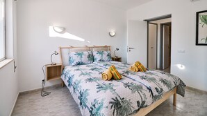 A bedroom with a bed, two bedside tables, a nightstand, a wall-mounted lamp, and a door leading to another room. - Apartment Casa Alegria (Carvoeiro)