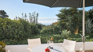 A round white table with two chairs, a bottle of wine, and two wine glasses on a patio with a view of lush greenery and a clear sky. - Apartment Casa Alegria (Carvoeiro)