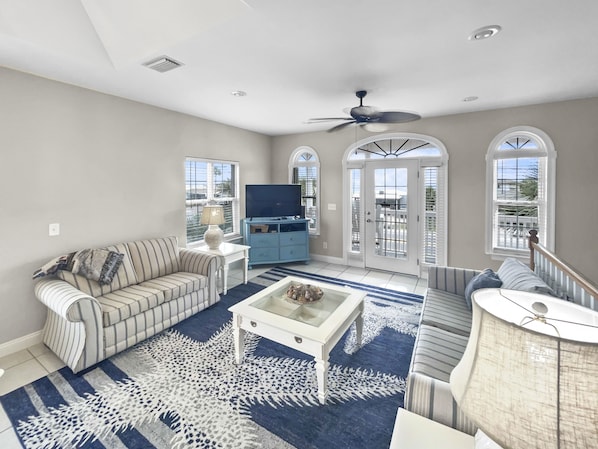 A spacious living room with a large sofa, a coffee table, and a TV. There are arched windows and a ceiling fan.