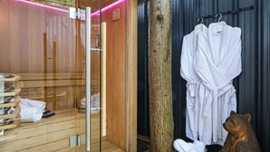 A sauna room with wooden flooring, a glass door, and a wooden tree trunk. There are white towels and a white robe hanging on a rack.