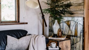 Una habitación acogedora con un sofá de cuero negro, una mesita de madera con una lámpara, un jarrón con una planta y un cuadro en la pared.