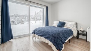 A bedroom with a large window, a bed with a blue blanket, and a bedside table with a lamp. - Modern chalet B (Gatineau)