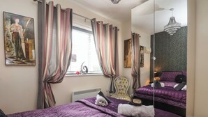 Ein Schlafzimmer mit einem großen Bett, einem Sessel, einem Fenster mit Vorhängen, einem Heizkörper und einem Spiegel, der das Bett und eine Deckenleuchte reflektiert.