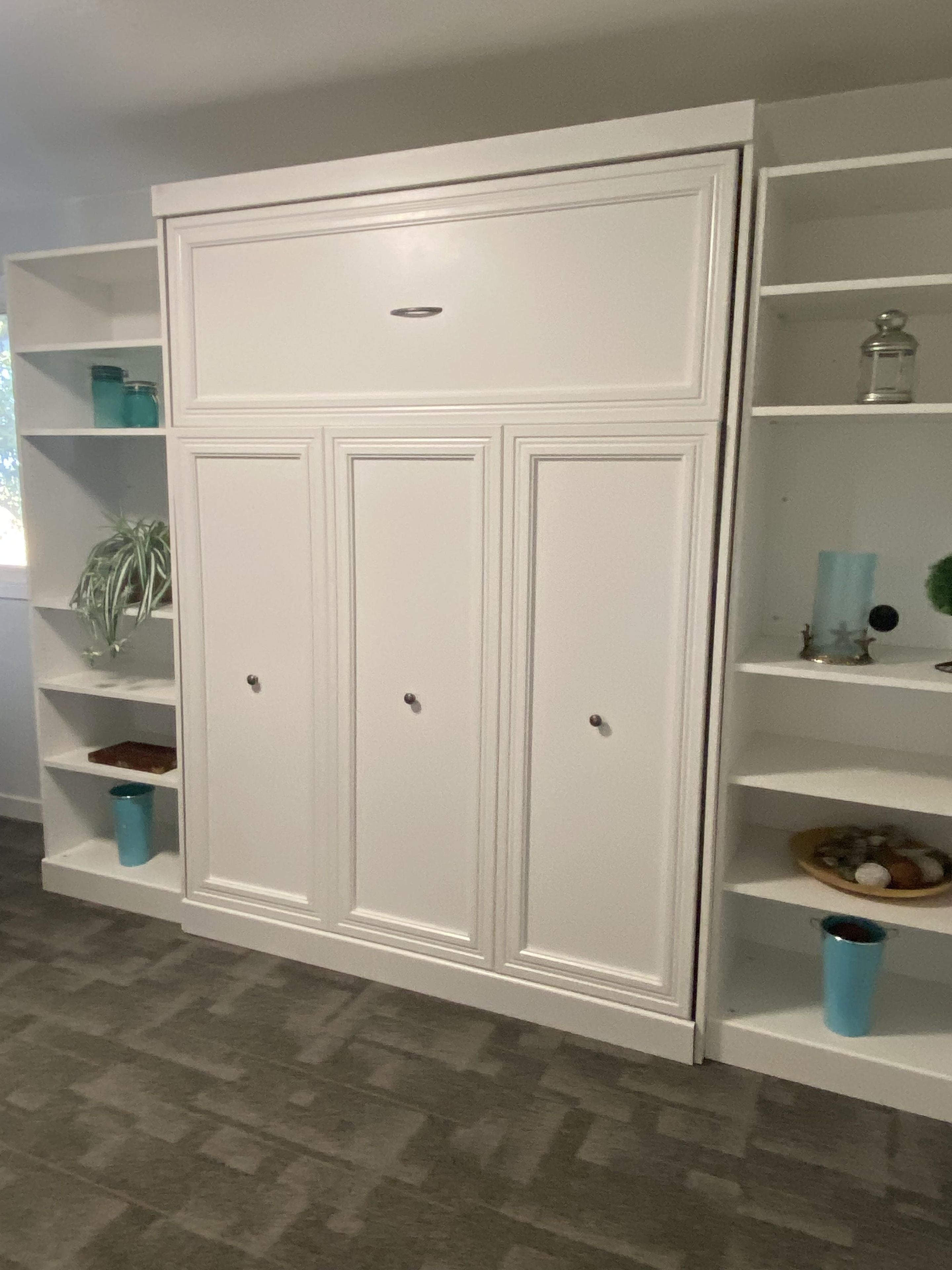 A white wardrobe with three doors and a small drawer, flanked by white shelving units with various items.