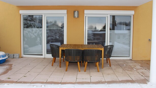 A patio with a wooden table and four chairs, a blue trash bin, and a sliding glass door.