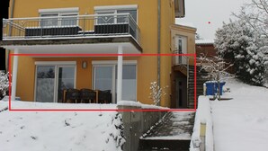 A yellow building with a balcony, steps leading up to the entrance, and snow on the ground.