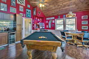 Een pooltafel in een ruimte met houten vloeren, een plafondventilator en een bar met een koelkast en planken.