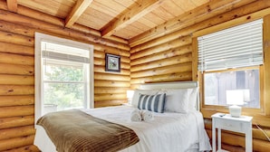 Une chambre située dans un chalet en rondins, avec un grand lit, une table de chevet et une fenêtre avec des stores.