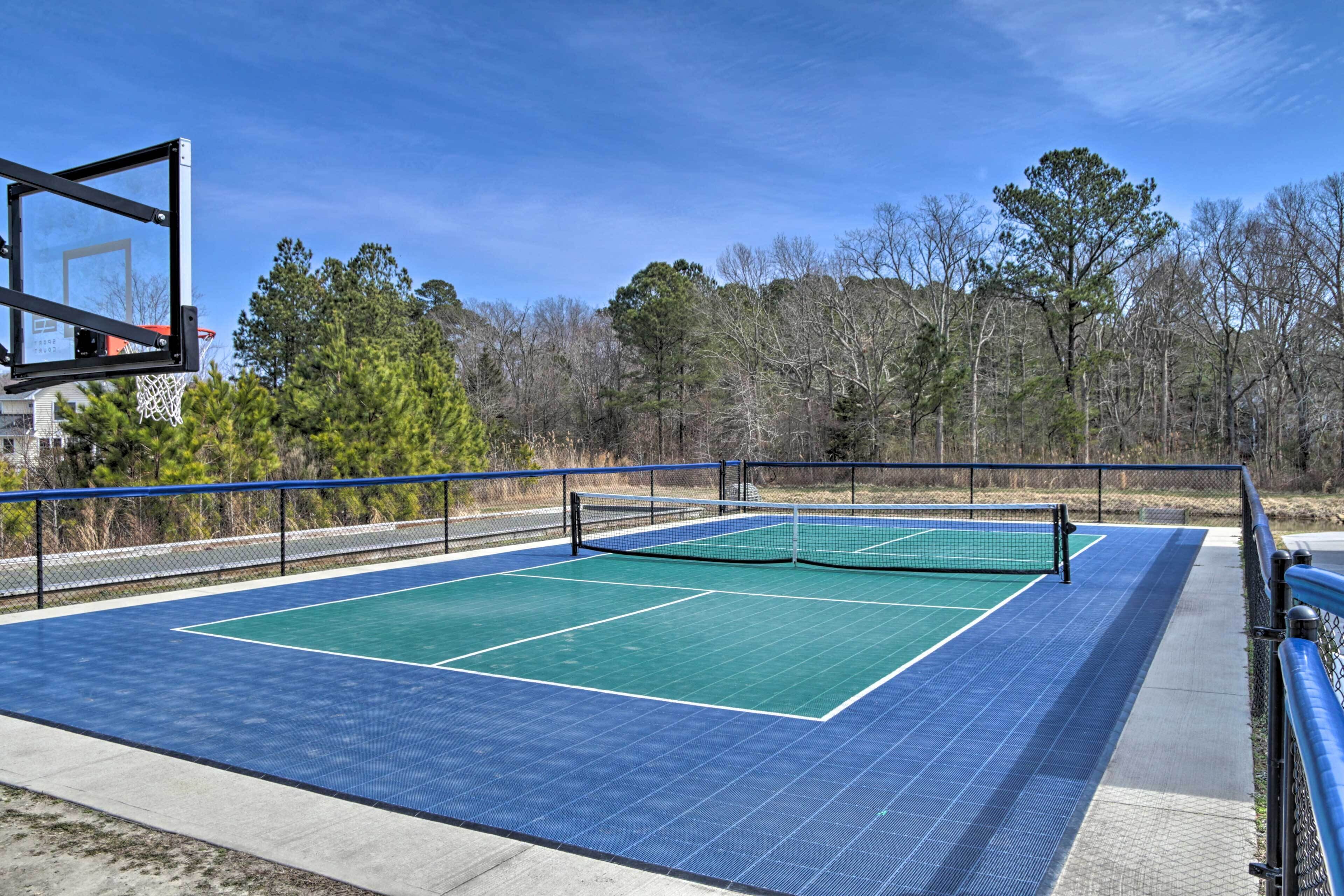 An outdoor tennis court with a basketball hoop, surrounded by trees and a clear sky.