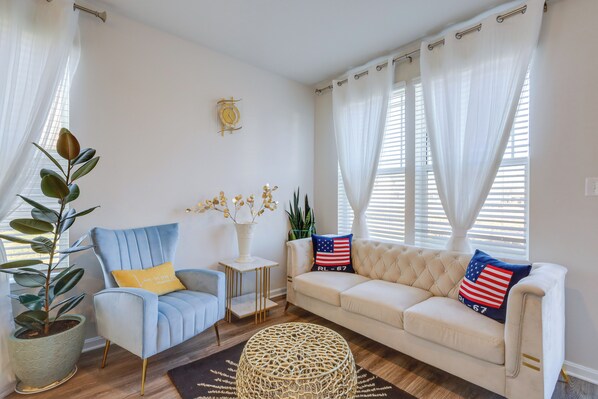 A living room with a blue armchair, a white sofa, and a coffee table. There are potted plants and a decorative wall clock. - 3 Mi to Bethany Beach: 'The Chief’s Quarters' Home (Ocean View)