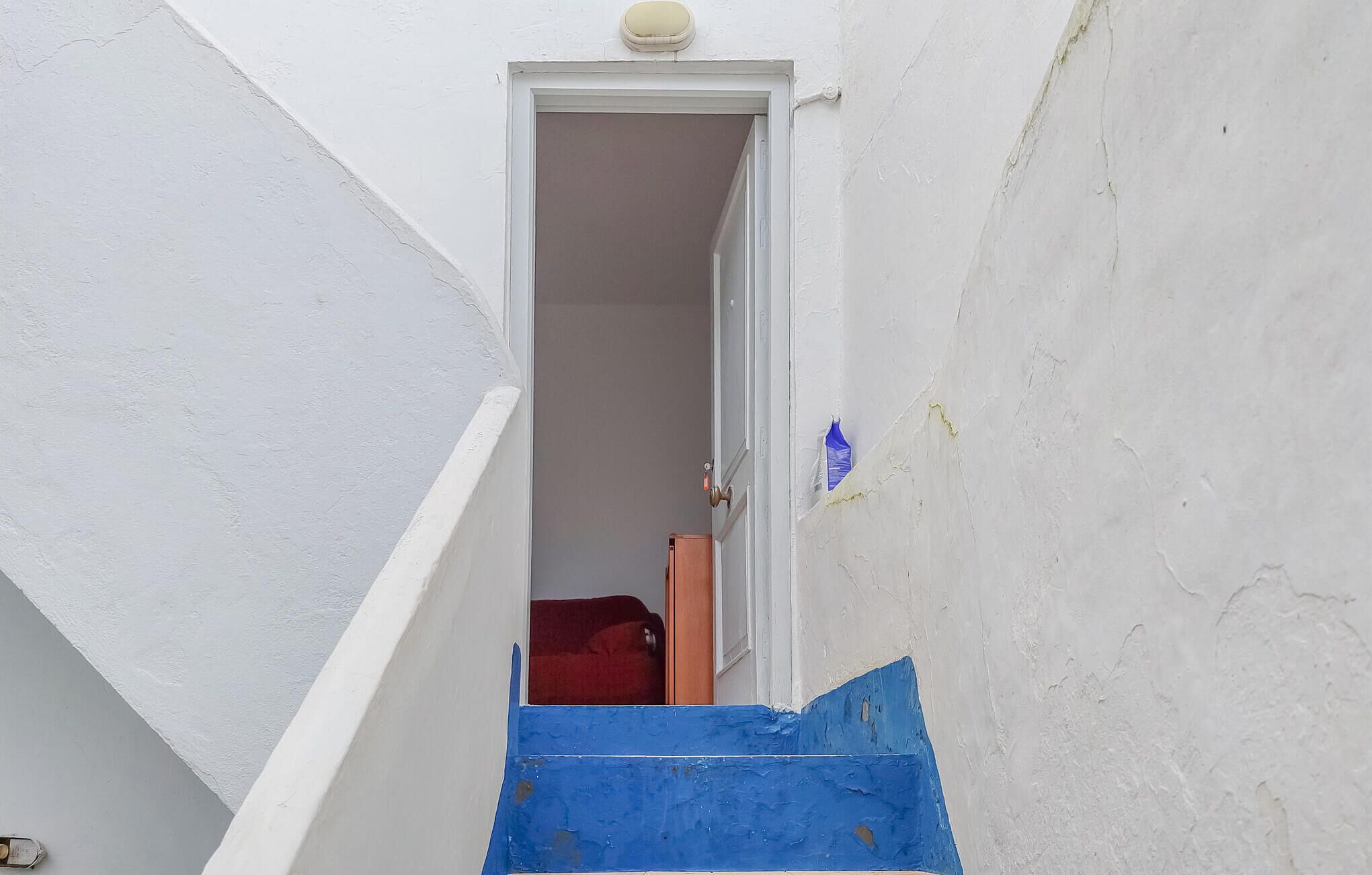 Un escalier étroit menant à une pièce où se trouvent une valise rouge et une porte.