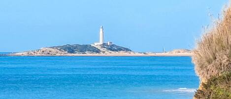 Un paisaje costero con un faro en una pequeña isla, agua azul cristalina y una costa rocosa.