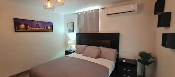 A bedroom with a bed, nightstands, a lamp, an air conditioner, and a wall-mounted TV.