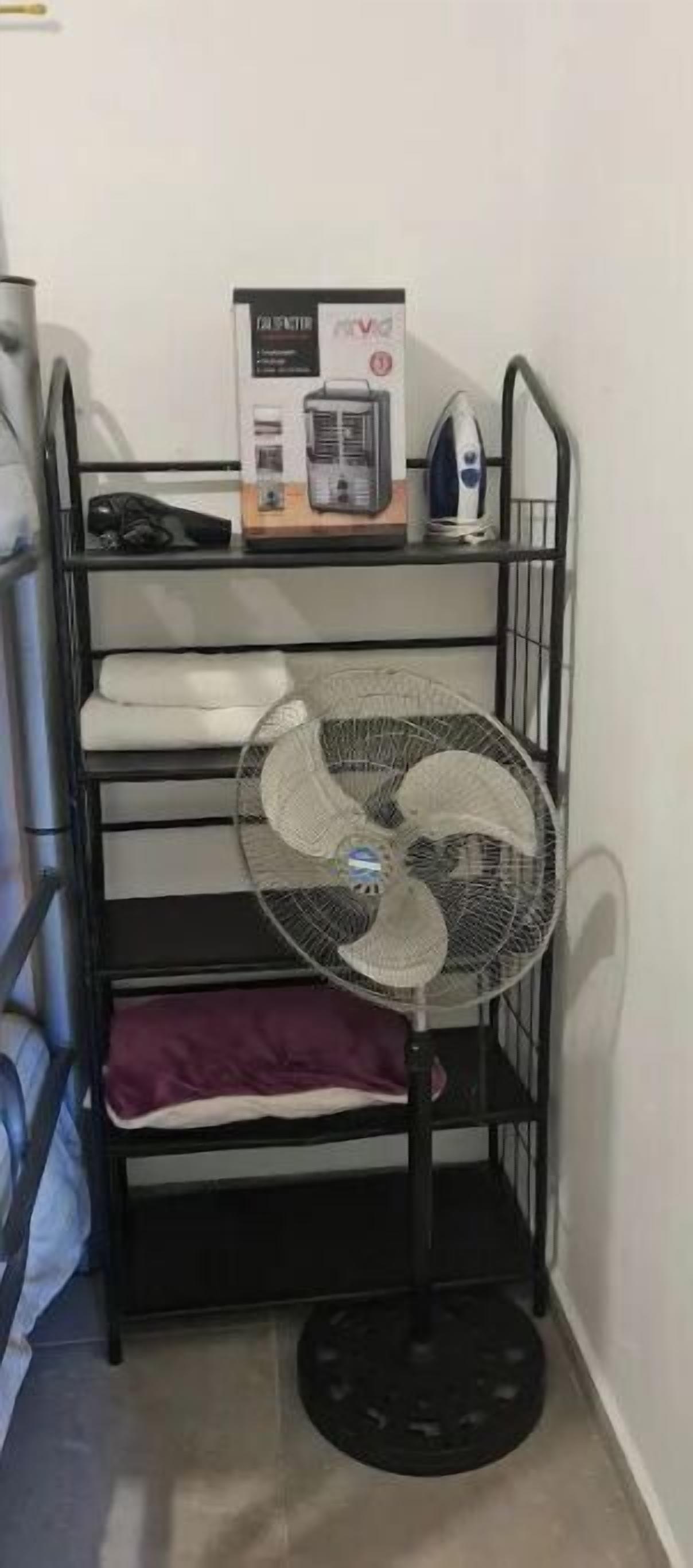 Una cama litera con un ventilador de pie en la esquina, una caja de detergente para ropa y una tetera en la parte superior.