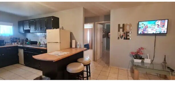 A kitchen with dark cabinets, a wooden counter, and two stools. There is a TV mounted on the wall and a glass cabinet with decorative items. - Gated and secured Appartment 6 mins from the beach (Puerto Peñasco)