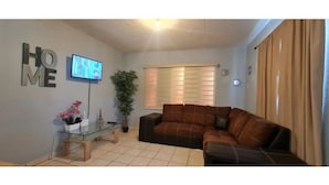 A living room with a brown leather sofa, a glass coffee table, a potted plant, and a TV mounted on the wall. - Gated and secured Appartment 6 mins from the beach (Puerto Peñasco)