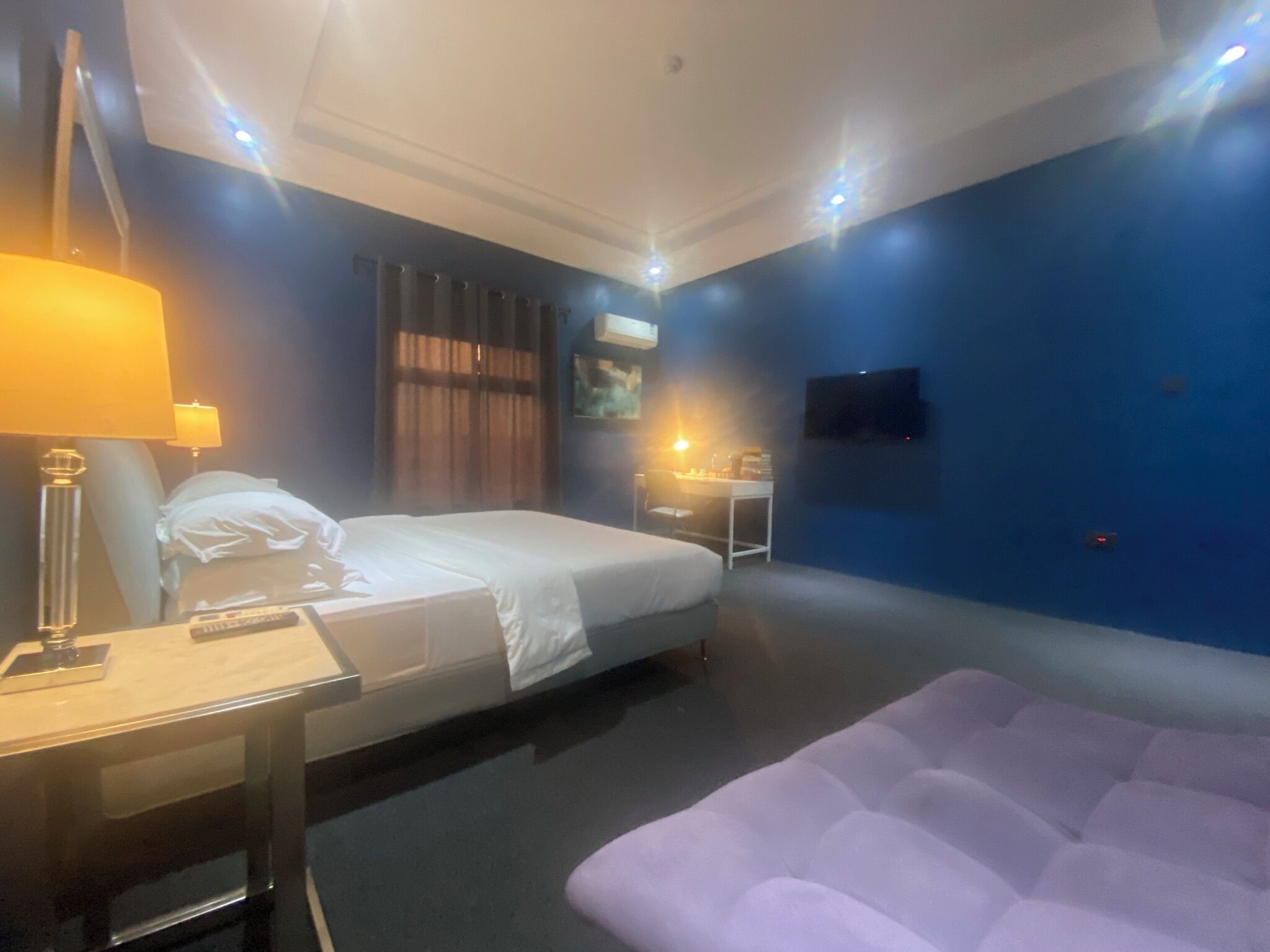 A hotel room with a bed, a desk with a lamp, a chair, a small table, and a TV mounted on the wall.