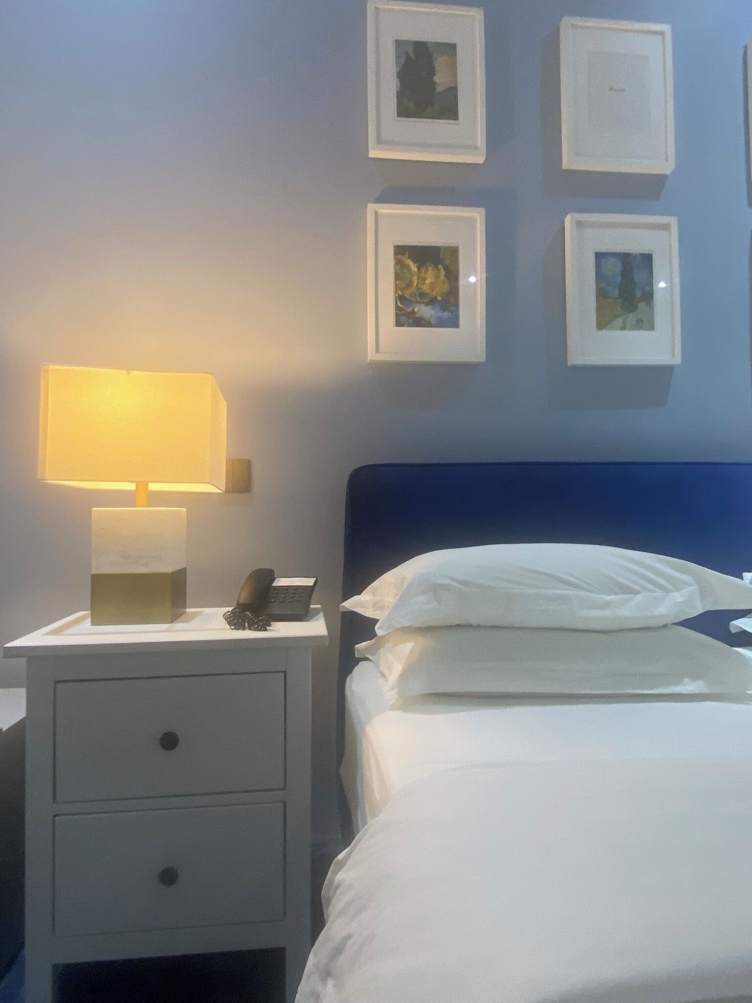 A hotel room with a bed, a nightstand with a lamp, a phone, and a drawer. There are four framed pictures on the wall.