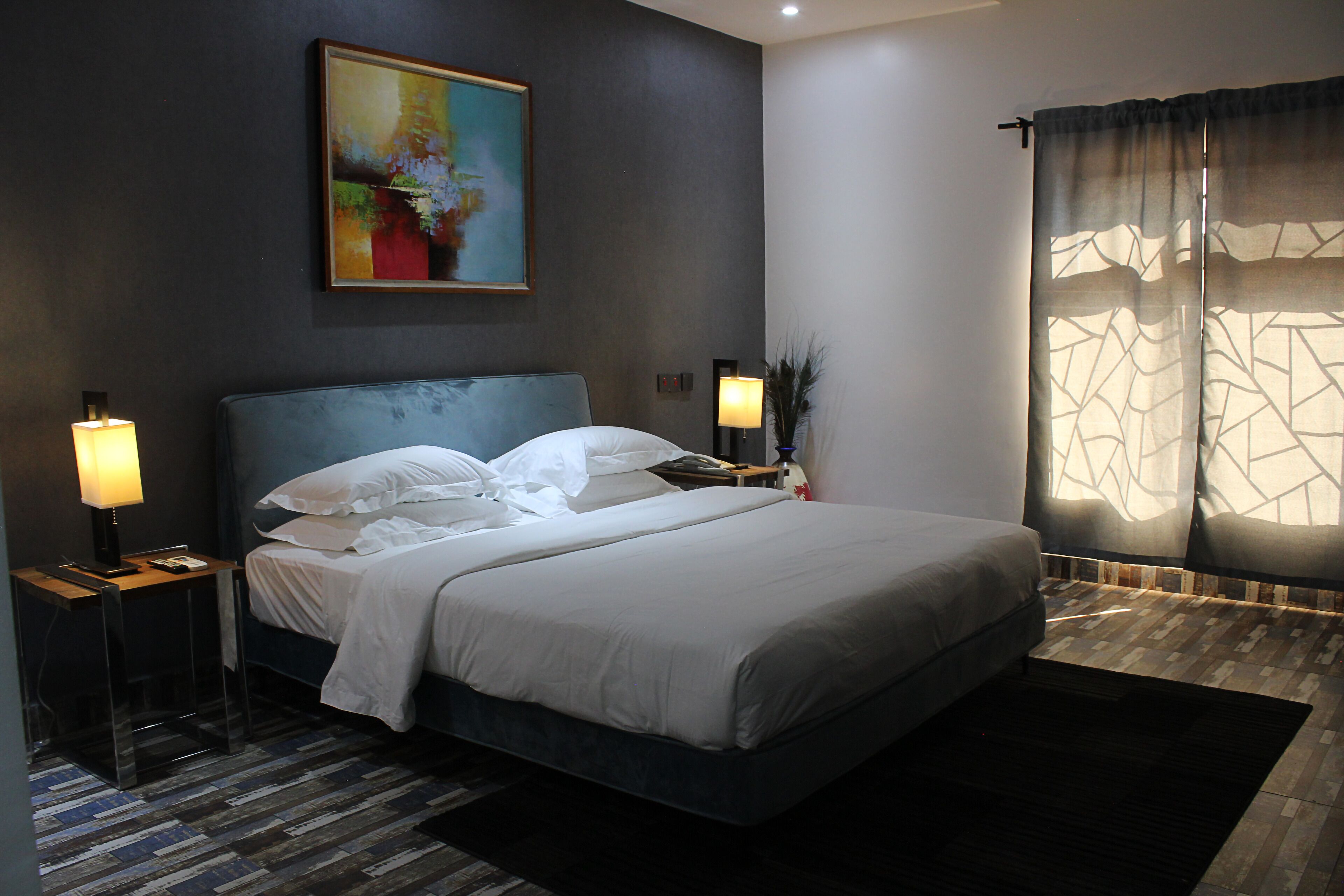 Una habitación de hotel moderna con una cama grande, dos mesitas de noche con lámparas, un cuadro en la pared y una ventana con cortinas estampadas.