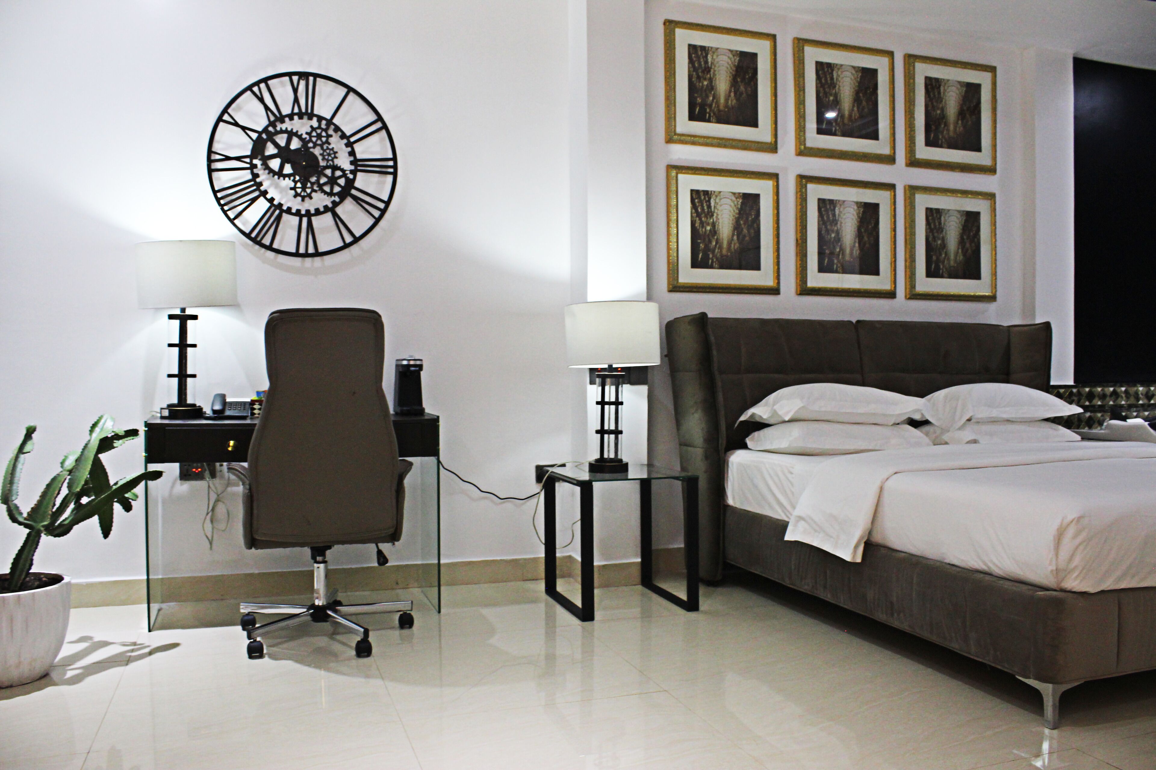 Ein Hotelzimmer mit einem Bett, einem Schreibtisch, einem Stuhl, einer Uhr und gerahmten Bildern an der Wand.