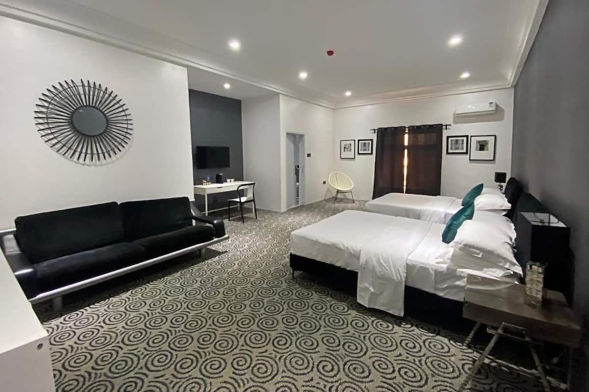 Ein Hotelzimmer mit zwei Betten, einem schwarzen Sofa, einem Schreibtisch mit Stuhl, einem Fernseher und einem gemusterten Teppich.