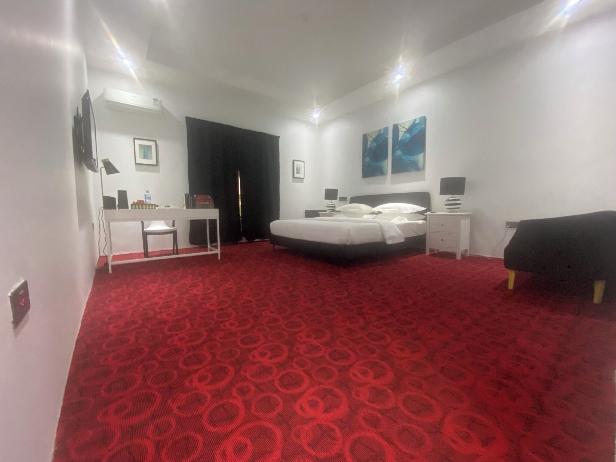 A hotel room with a red patterned carpet, a bed, a desk, a chair, a TV, and artwork on the walls.