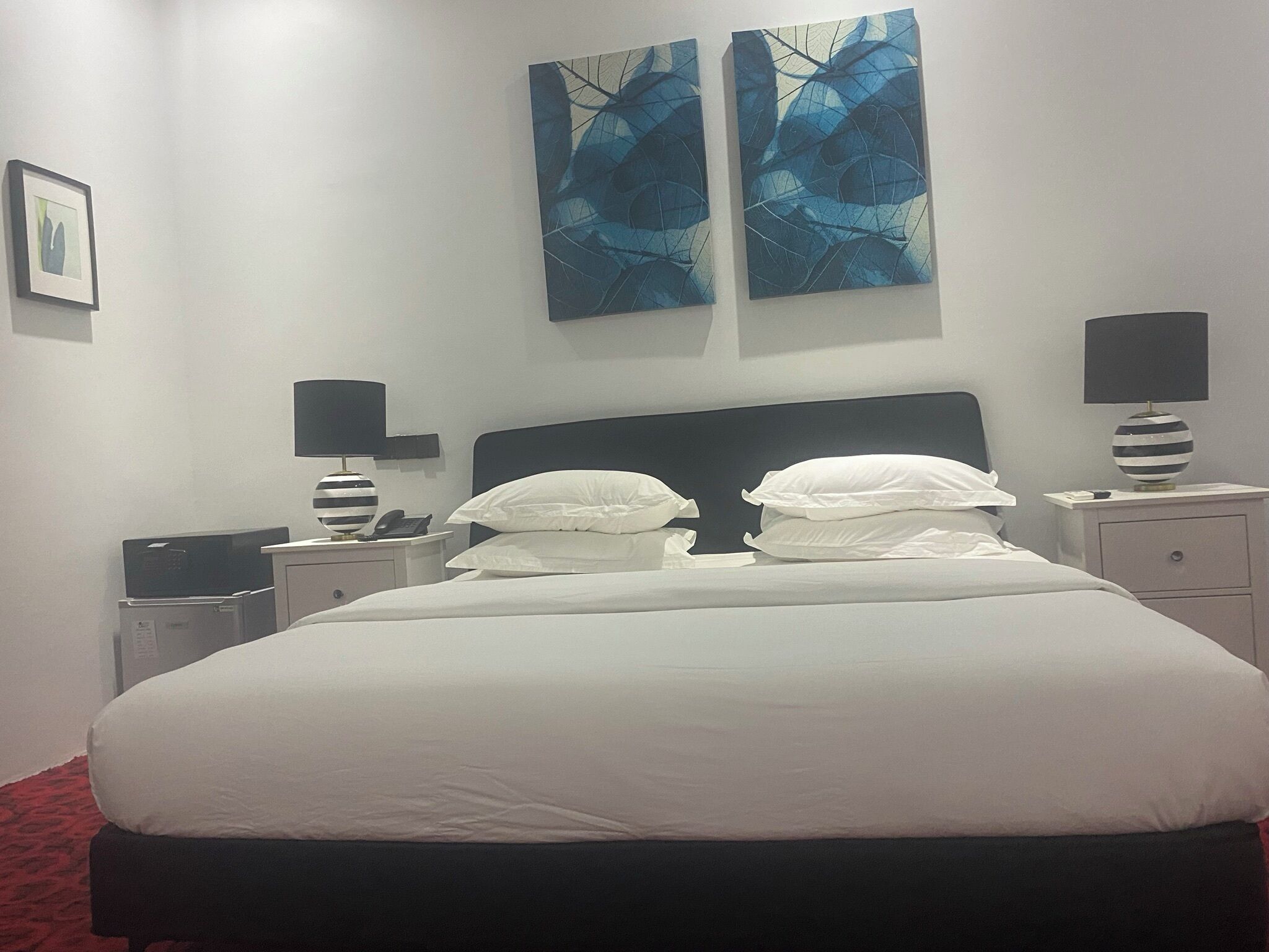 Una cama bien hecha con sábanas blancas, dos mesitas de noche con lámparas y dos cuadros abstractos azules en la pared.