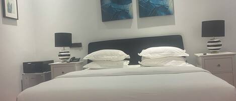 Una cama bien hecha con sábanas blancas, dos mesitas de noche con lámparas y dos cuadros abstractos azules en la pared.