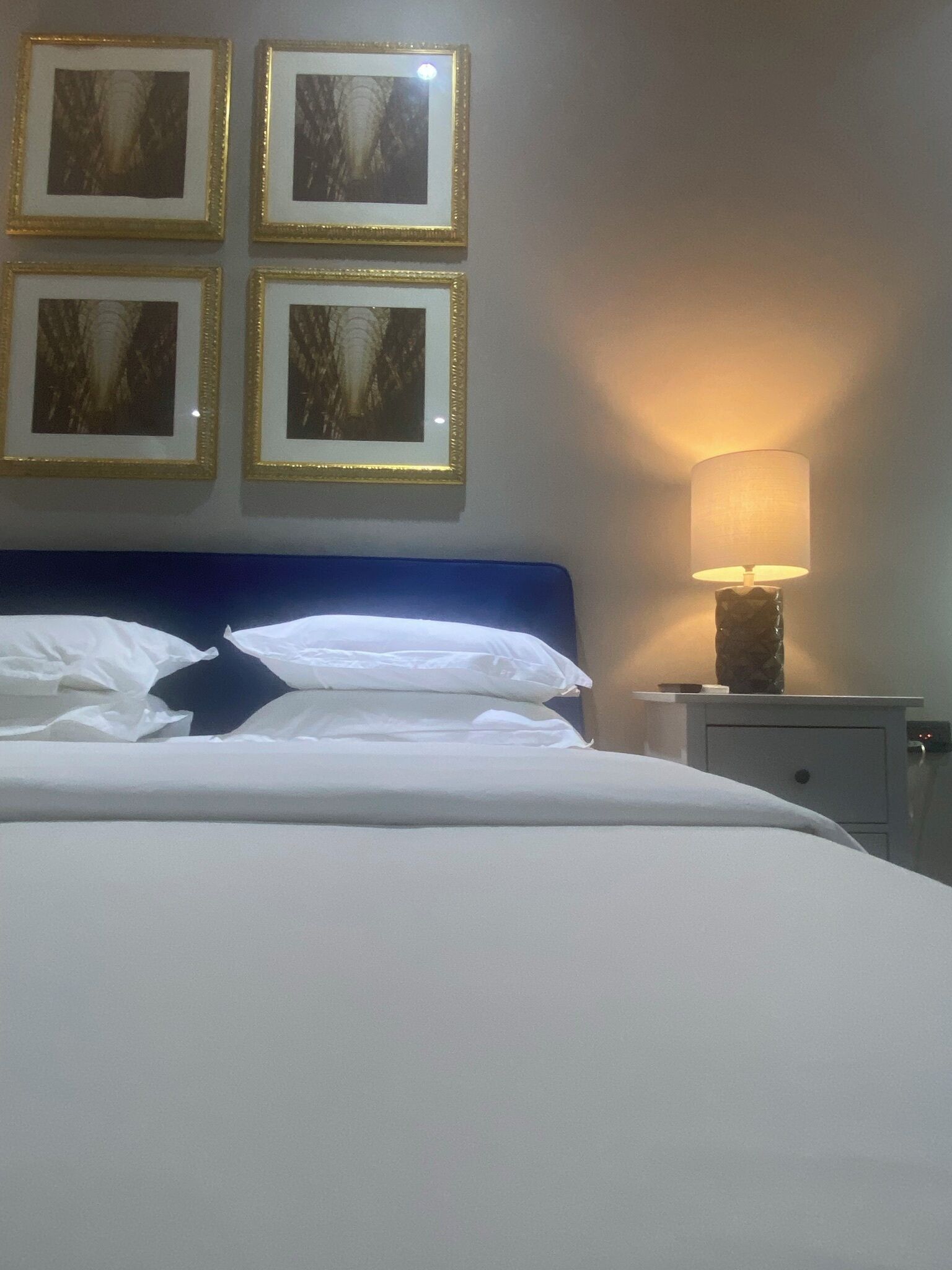 A neatly made bed with white linens and a blue headboard. There are four framed pictures on the wall above the bed. A bedside lamp is on the nightstand to the right.