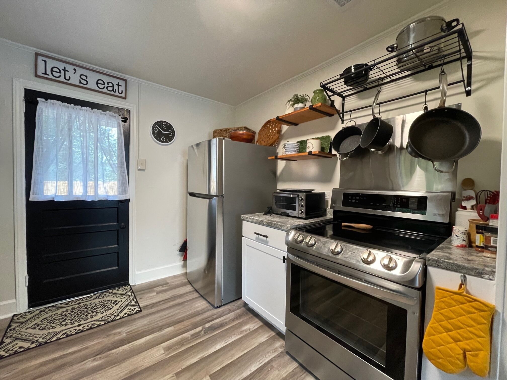 Een kleine keuken met een koelkast, magnetron, oven en broodrooster. Een klok en een bord met de tekst 'let's eat' zijn zichtbaar.