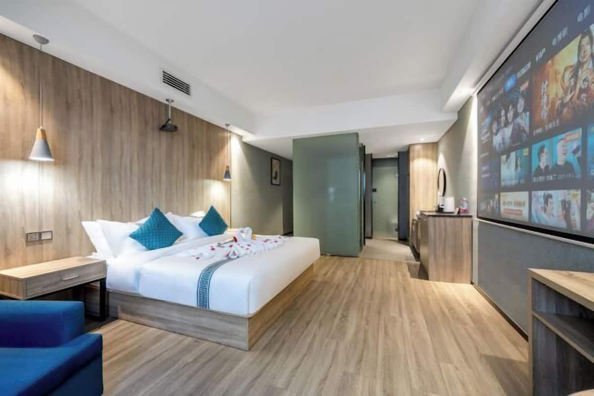 Une chambre d’hôtel moderne avec un plancher en bois, un lit recouvert de draps blancs et agrémenté d’oreillers turquoise, un bureau avec une lampe et un écran de télévision diffusant diverses chaînes.